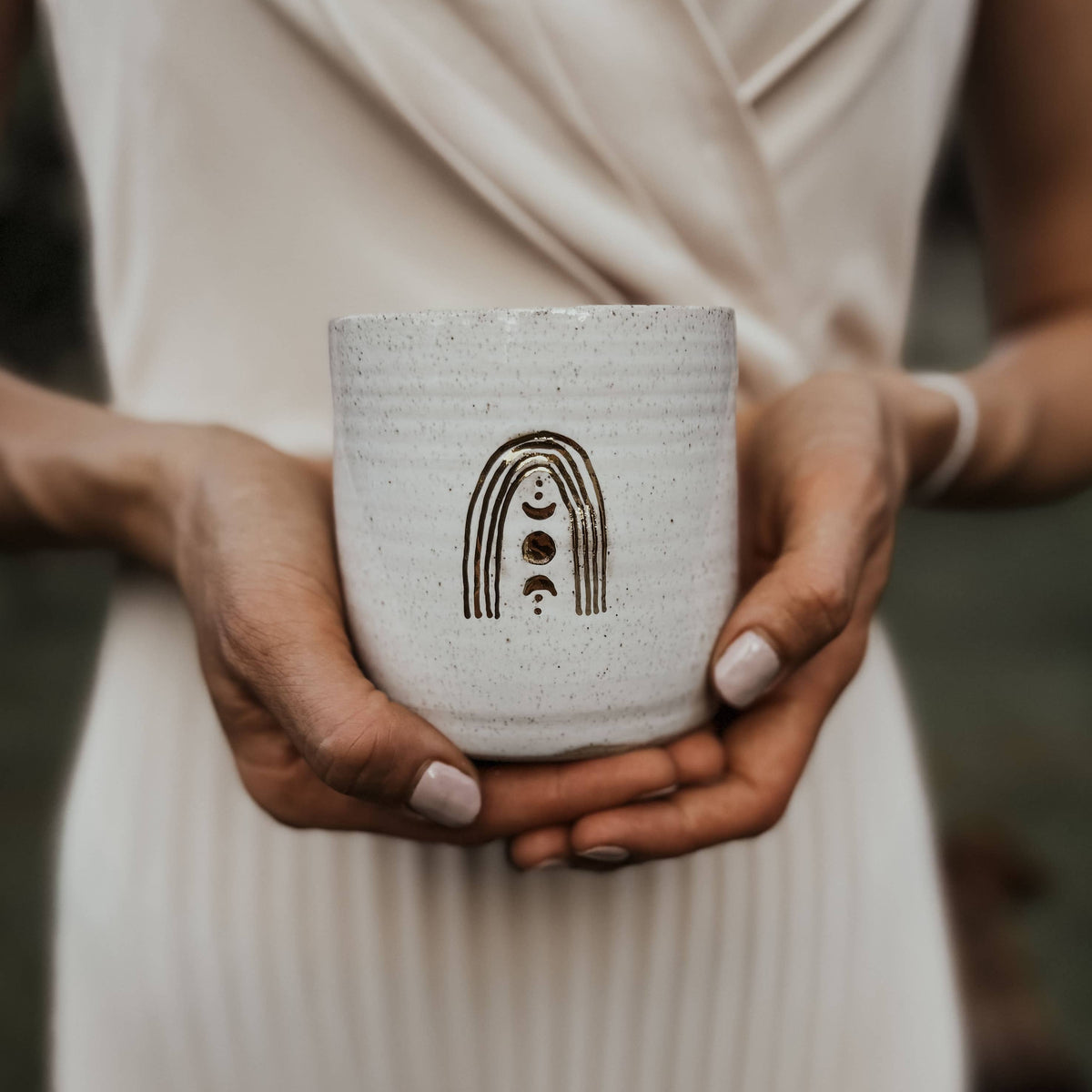 Person holding a white ceramic cup with a design, wearing a beige dress.