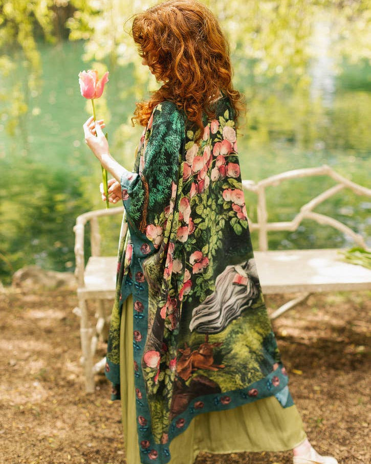 A woman stands outdoors by a lake holding a pink flower, wearing a dark green floral shawl over a light olive dress. A white bench and soft green foliage are in the background.