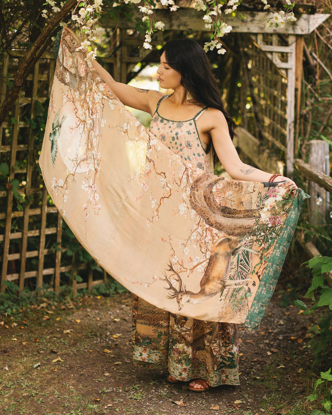 Woman in a floral dress holding a large, decorative fan in a garden setting.